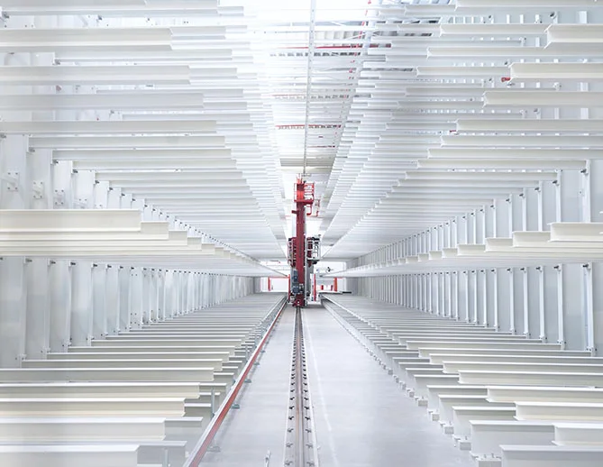 Moderne automatisierte Lagerhalle mit weißen Metallregalen und einem roten robotergesteuerten Kommissioniersystem, das entlang des schmalen Korridors zwischen den Regalen arbeitet.