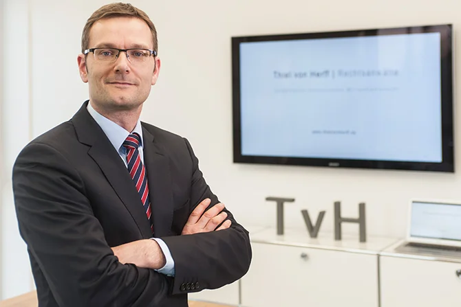 Ein professioneller Mann in einem Anzug mit Brille steht selbstbewusst in einer Büroumgebung, mit einem Präsentationsbildschirm und dem Firmenkürzel im Hintergrund.