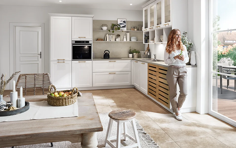 Helle Landhausküche mit Front CASCADA 774 in Weiß, kombiniert mit Holzdetails und goldenen Griffen, offene Gestaltung mit viel Tageslicht.