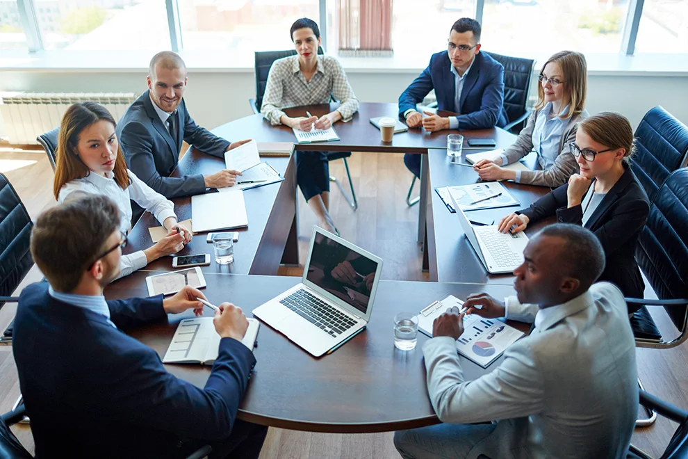 Eine vielfältige Gruppe von Fachleuten, die an einem Tisch mit Laptops und Dokumenten in einem modernen Büro an einer Besprechung teilnehmen und über Geschäftsstrategien diskutieren.