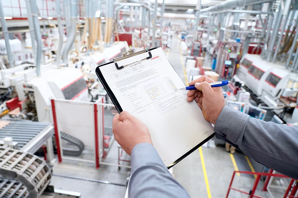 Blick über die Schulter einer Person, die eine Checkliste auf einem Klemmbrett in einem modernen industriellen Fertigungsbetrieb mit Maschinen im Hintergrund überprüft.