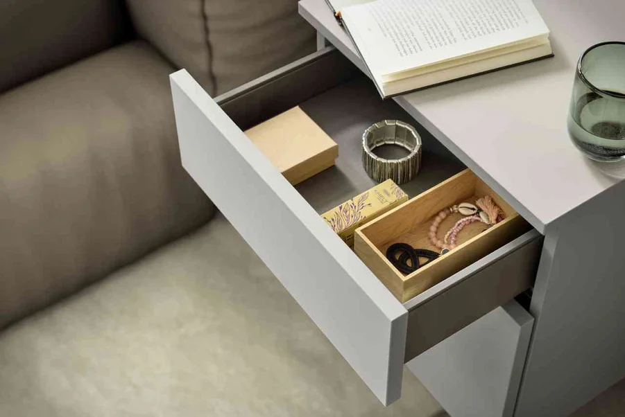 Offene Schublade mit Holzboxen, Schmuck und einem Glas auf einem modernen Tisch neben einer Couch.