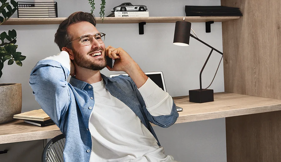 Ein zufriedener junger Berufstätiger unterhält sich auf seinem Smartphone, während er bequem in einem stilvollen Homeoffice mit minimalistischer Einrichtung und stimmungsvoller Beleuchtung sitzt.