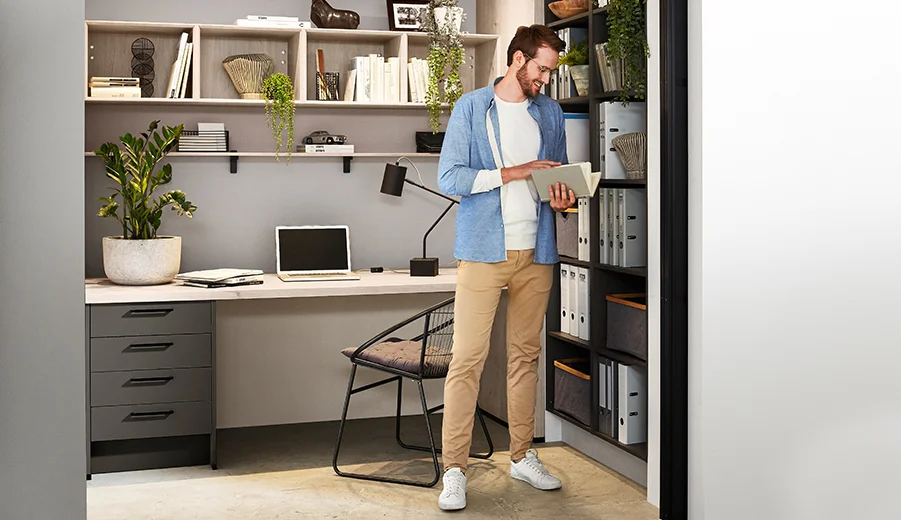Ein konzentrierter Einzelner in einem stilvollen Home-Office-Setup, der durch ein Buch an einem organisierten Regal blättert und eine Mischung aus Komfort und Produktivität widerspiegelt.